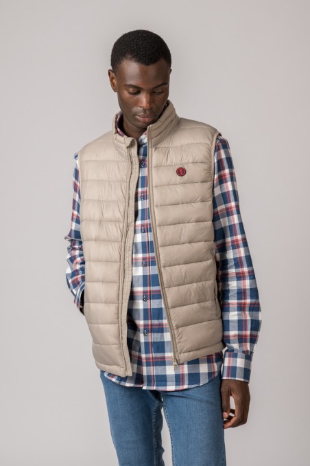 Homem a vestir colete bege acolchoado sobre camisa xadrez e calças de ganga.
