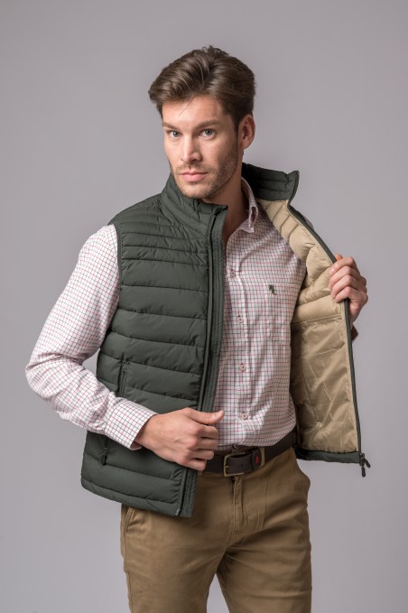 Homem veste colete almofadado verde escuro aberto mostrando interior bege, camisa branca com quadrados vermelhos e calças castanhas.