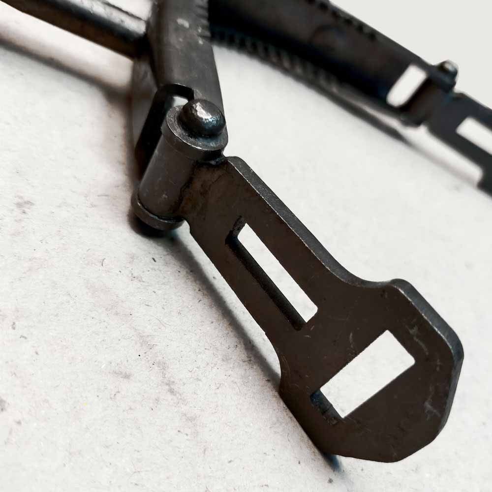 Peça metálica escura articulada com aberturas retangulares sobre superfície clara