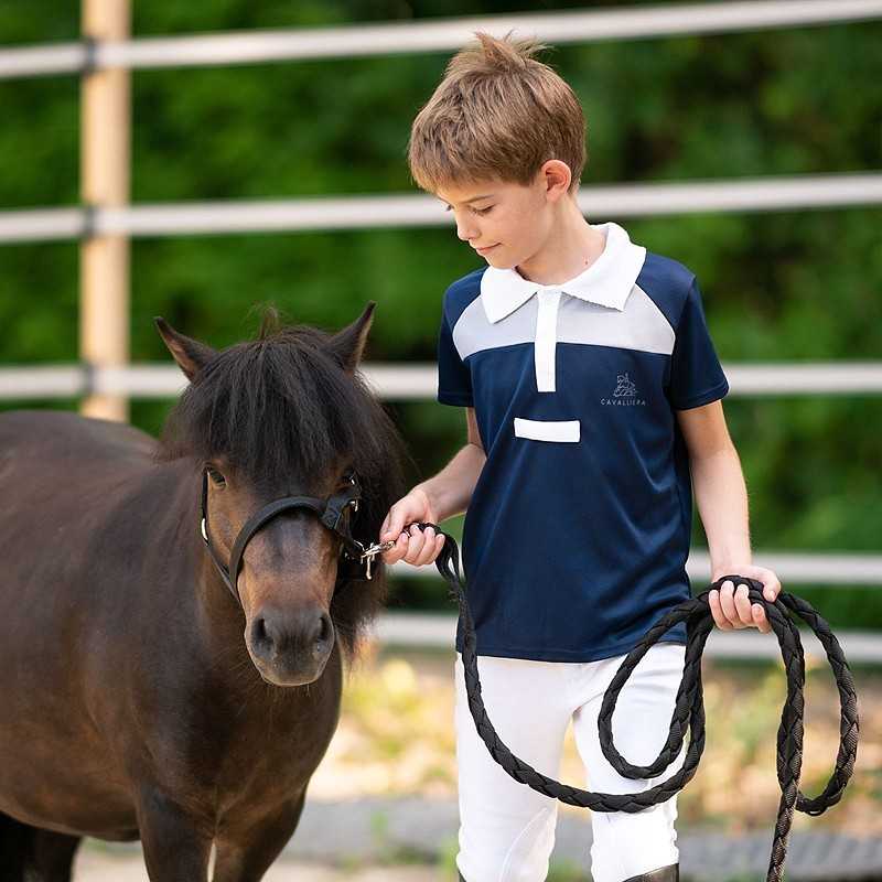 Menino com camisa polo azul escura com gola branca a segurar a rédea preta de um pônei castanho ao ar livre