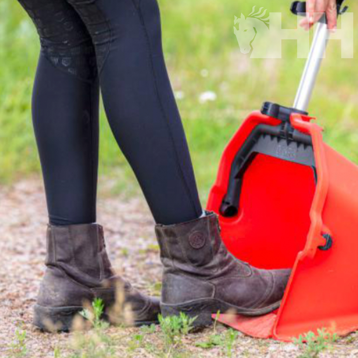 Pernas com leggings pretas e botas castanho escuro ao lado de uma pá vermelha com pega metálica em terra
