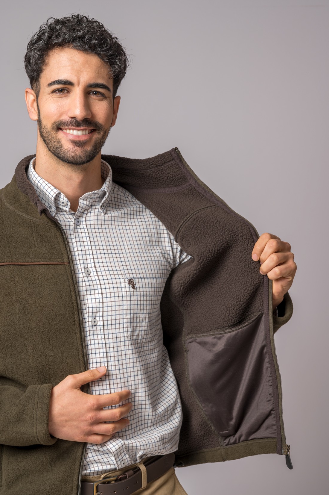 Homem com camisa branca quadriculada, casaco verde polar aberto mostrando interior castanho e bolso, cinto e calças bege.