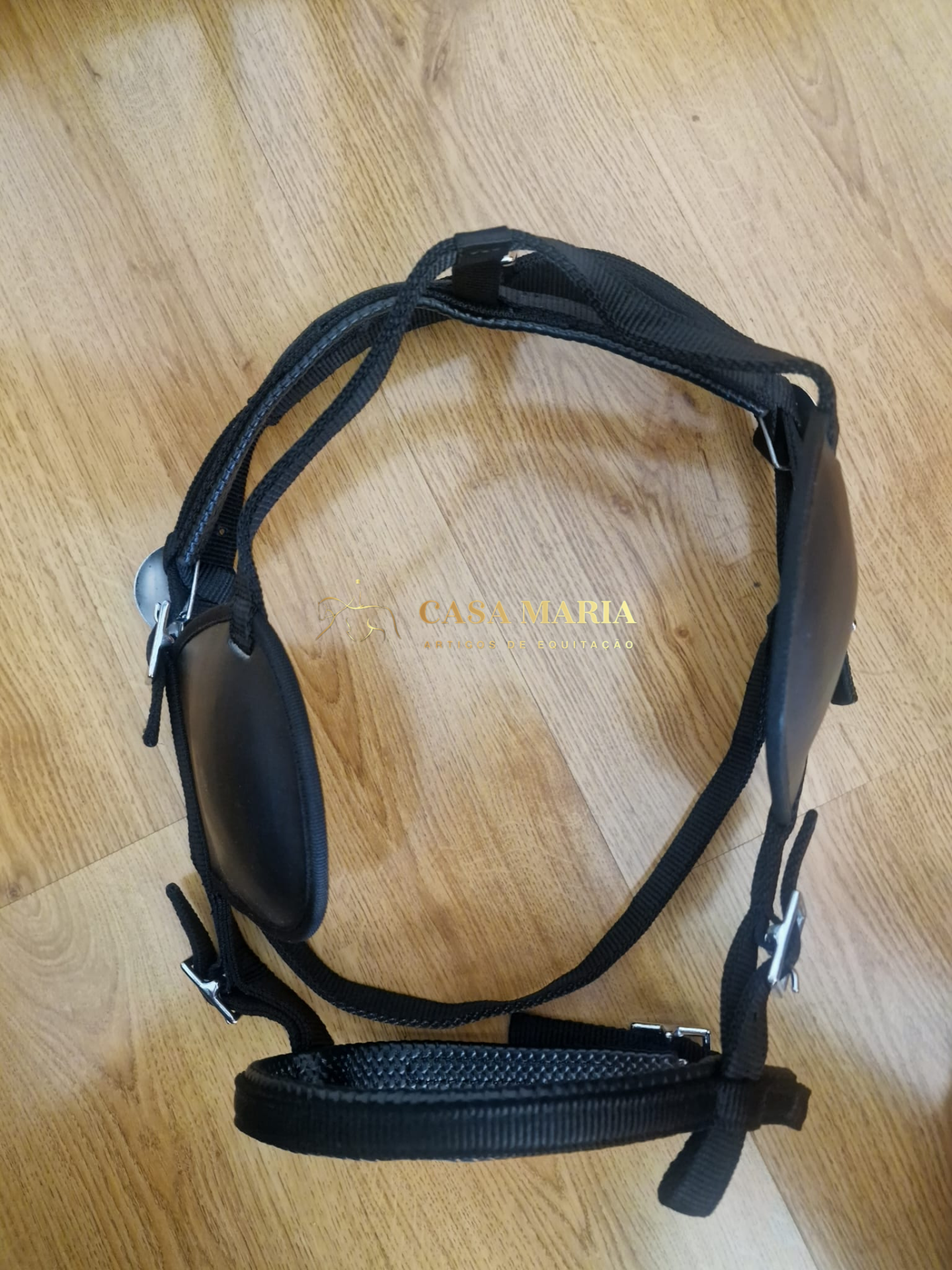 Arnês preto ajustável com acolchoamento e fivelas metálicas sobre chão de madeira