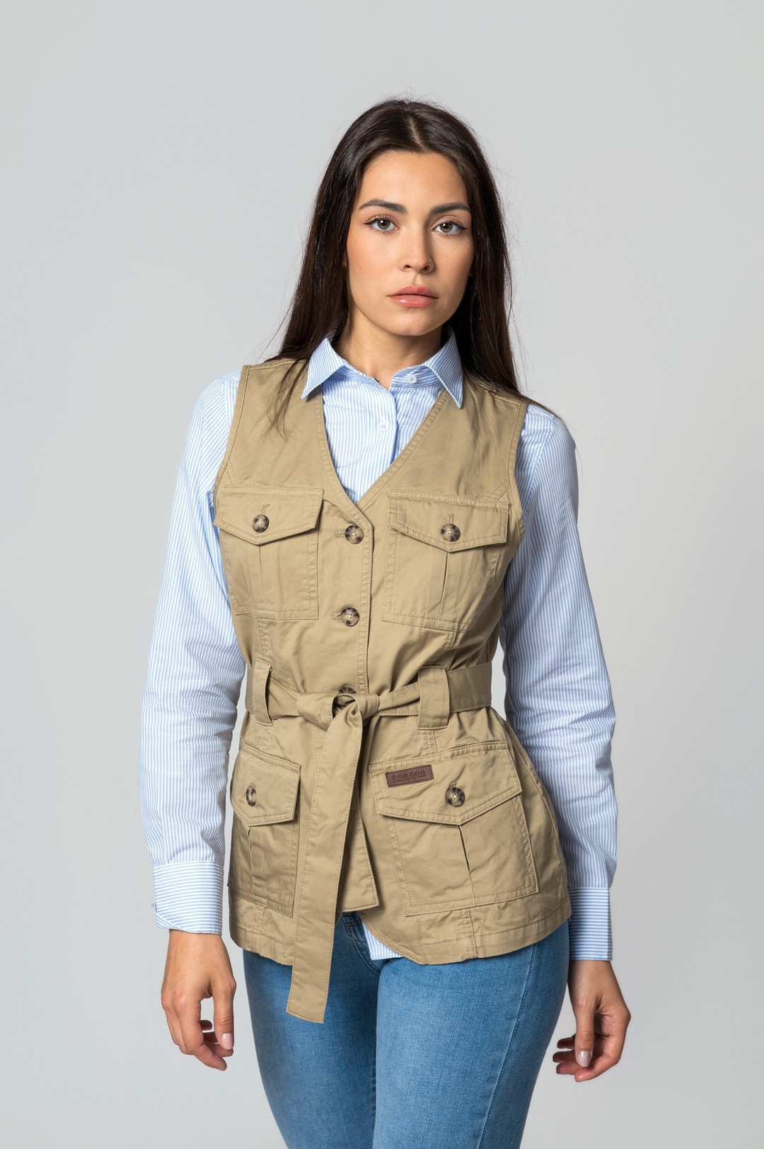 Mulher com colete bege de bolsos e cinto, camisa azul clara às riscas e jeans azuis.