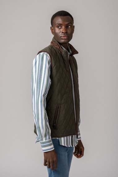 Homem a usar colete matelassê verde-oliva por cima de camisa riscada azul e branco, com jeans azul claro.