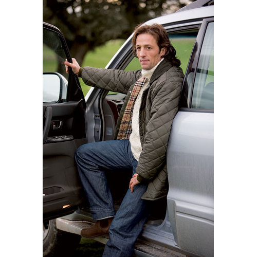 Homem sentado na soleira de carro prateado com casaco acolchoado cinza e jeans