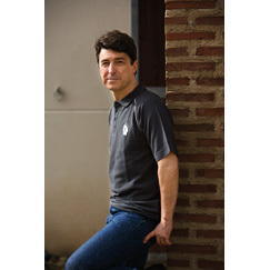 Homem com camisa preta e calças azuis encostado numa parede de tijolos vermelhos