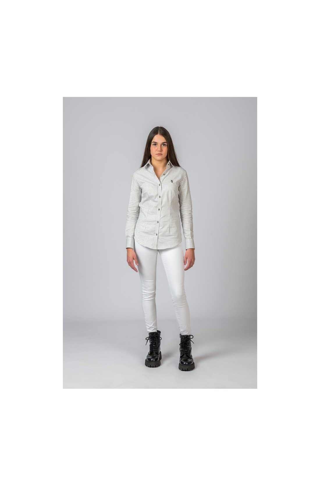 Mulher com camisa xadrez cinza clara, calças brancas e botas pretas em fundo cinza