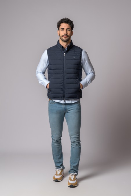 Homem com colete azul escuro, camisa azul clara, jeans e ténis bege