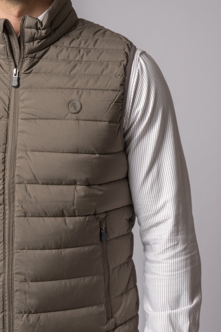 Colete acolchoado bege castanho com fecho zip e bolso lateral, usado sobre camisola branca de malha