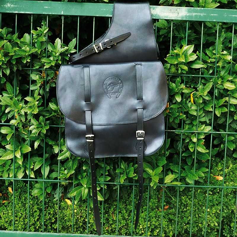 Mochila preta de couro com fivelas metálicas pendurada numa vedação verde com plantas ao fundo