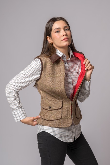 Mulher vestindo colete castanho em tweed aberto mostrando interior vermelho, camisa branca xadrez preta e calças pretas com fundo cinza