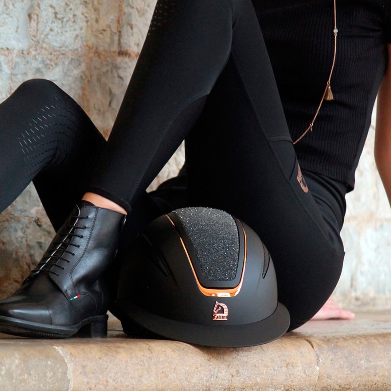 Capacete de equitação preto matte com detalhes laranja e bota de couro preta ao lado, pessoa sentada em degrau de pedra