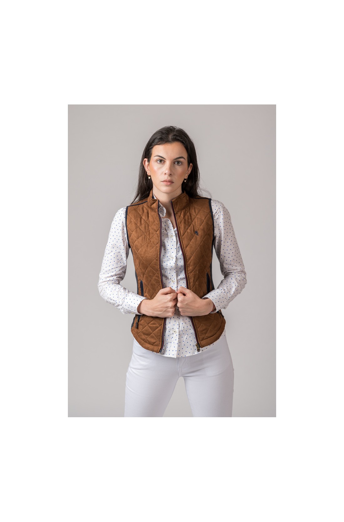 Mulher a usar colete matelassé castanho sobre camisa branca com padrão de pontos pretos e calças brancas