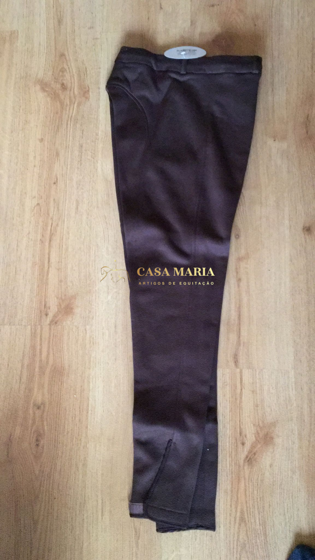 Calças castanhas escuras sobre chão de madeira clara com etiqueta branca na cintura