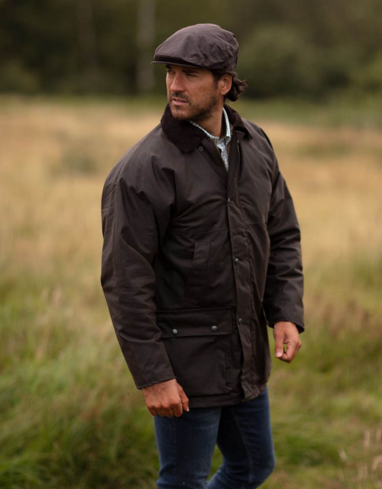 Homem com casaco castanho, calças azuis e boina castanha em campo com relva