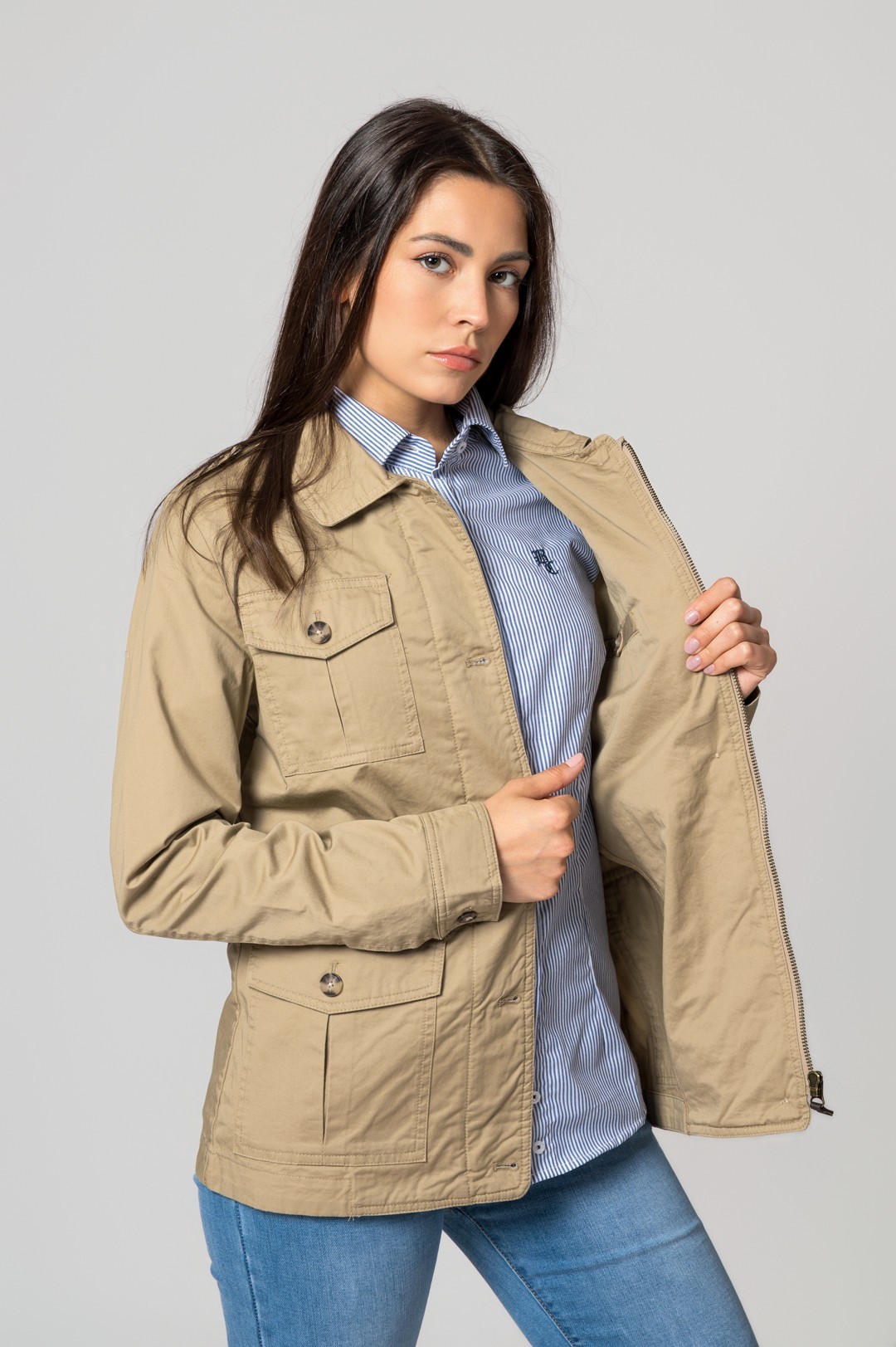 Mulher com blusão bege, camisa riscada azul e branca, calças de ganga, fundo cinza