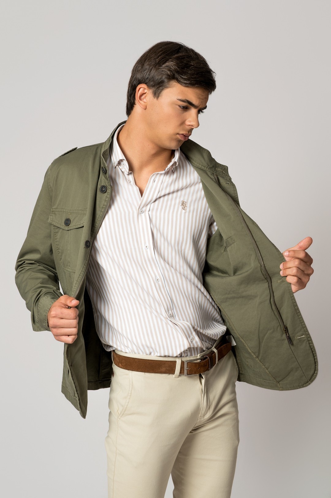 Modelo a vestir camisa riscada branca e bege, casaco verde militar aberto, calças bege claras e cinto castanho.