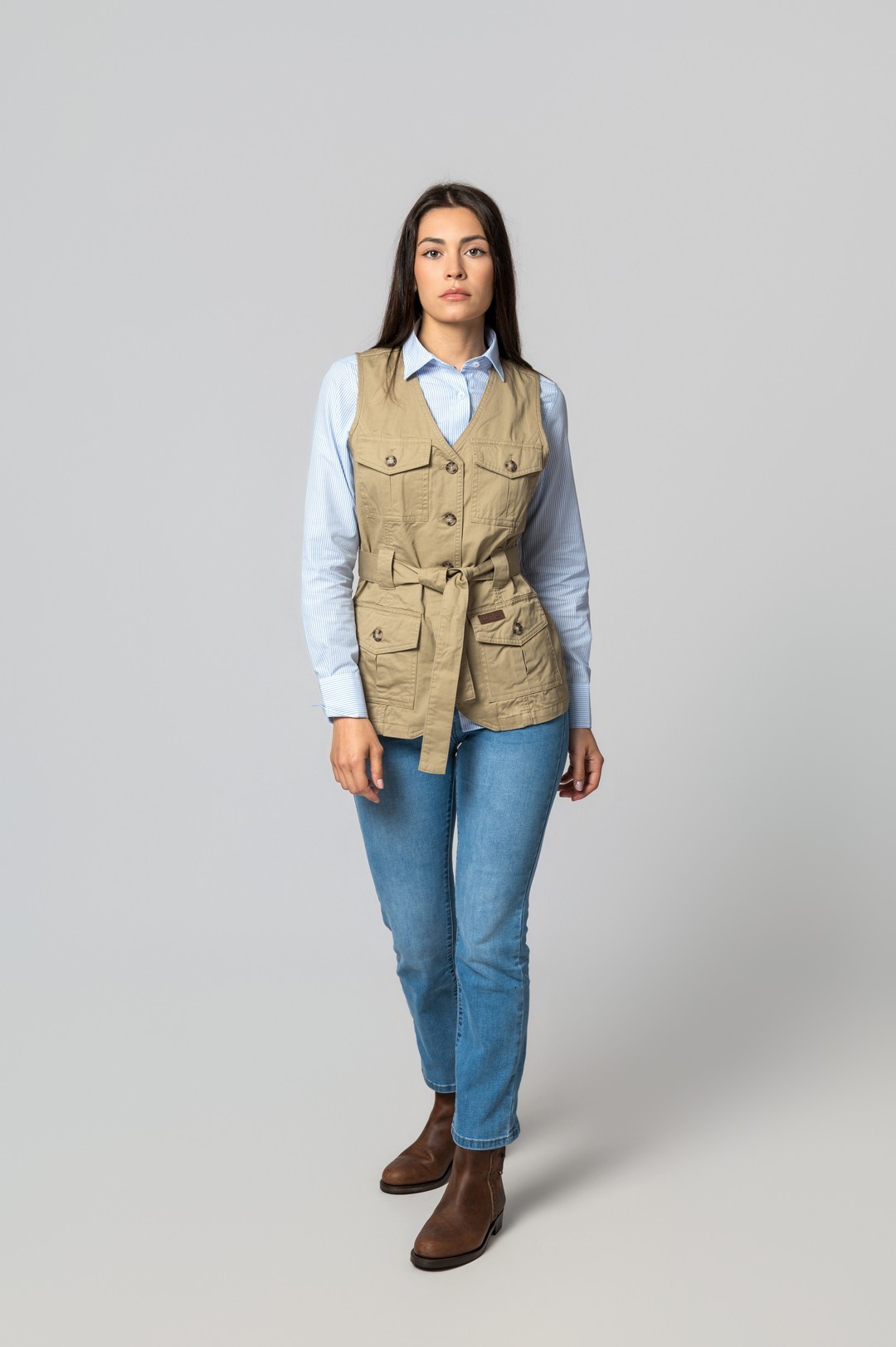 Mulher com colete bege, camisa azul de riscas, jeans azul claro e botas castanhas