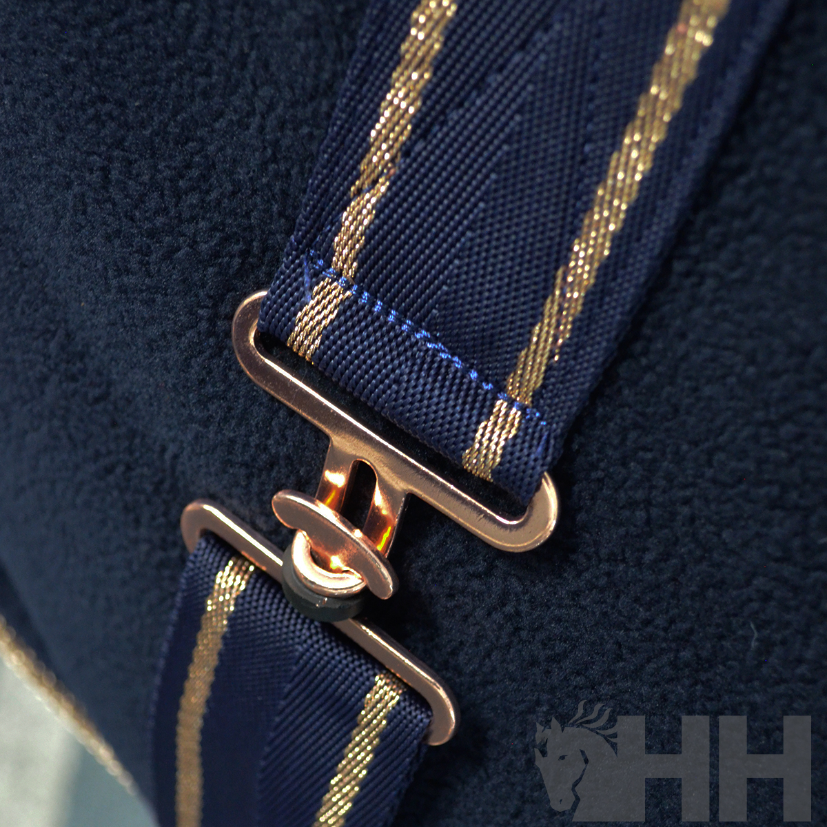 Alça azul com riscas douradas e fivela metálica sobre tecido azul escuro com logotipo HH