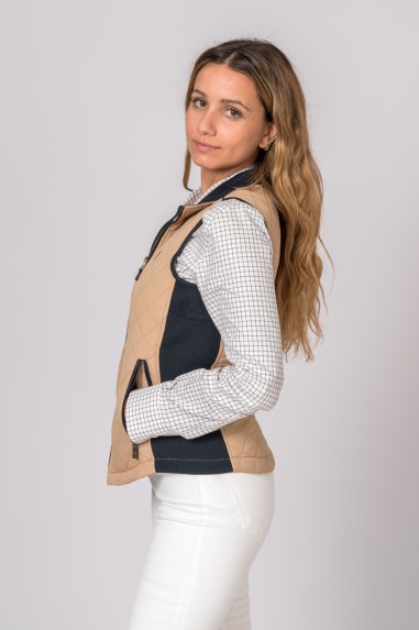 Mulher de perfil a usar colete bege matelassê com detalhes em azul e camisa branca xadrez