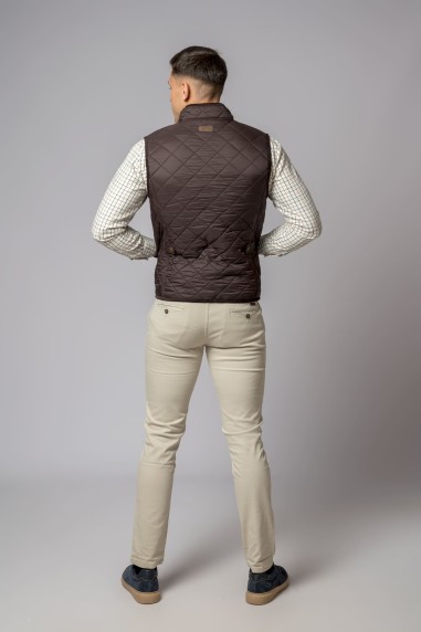 Homem de costas com colete acolchoado castanho, camisa branca xadrez, calças bege e sapatos escuros