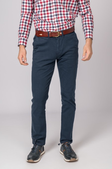 Homem com calças azuis, cinto castanho, camisa xadrez e sapatos pretos em fundo cinza