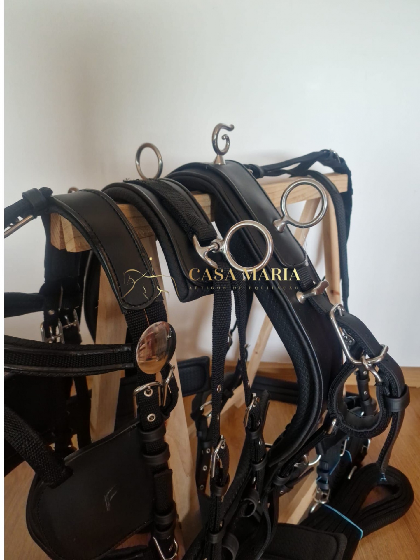 Arreios de couro preto com acessórios metálicos pendurados num suporte de madeira