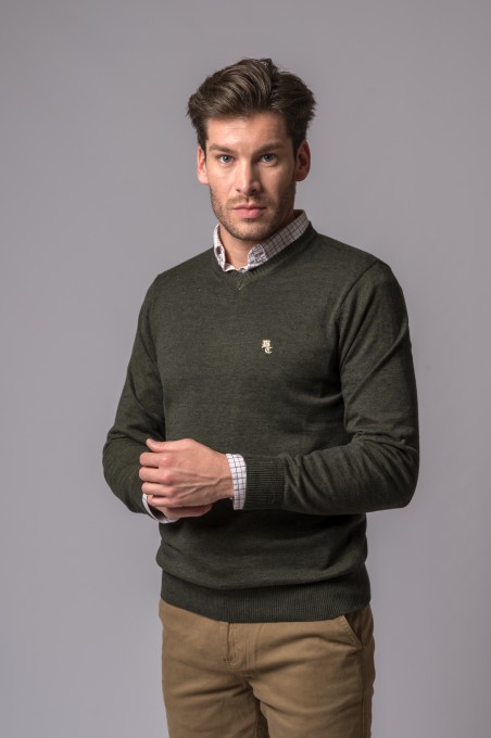 Homem a vestir camisola verde escuro de malha com gola em V sobre camisa xadrez clara e calças castanhas.