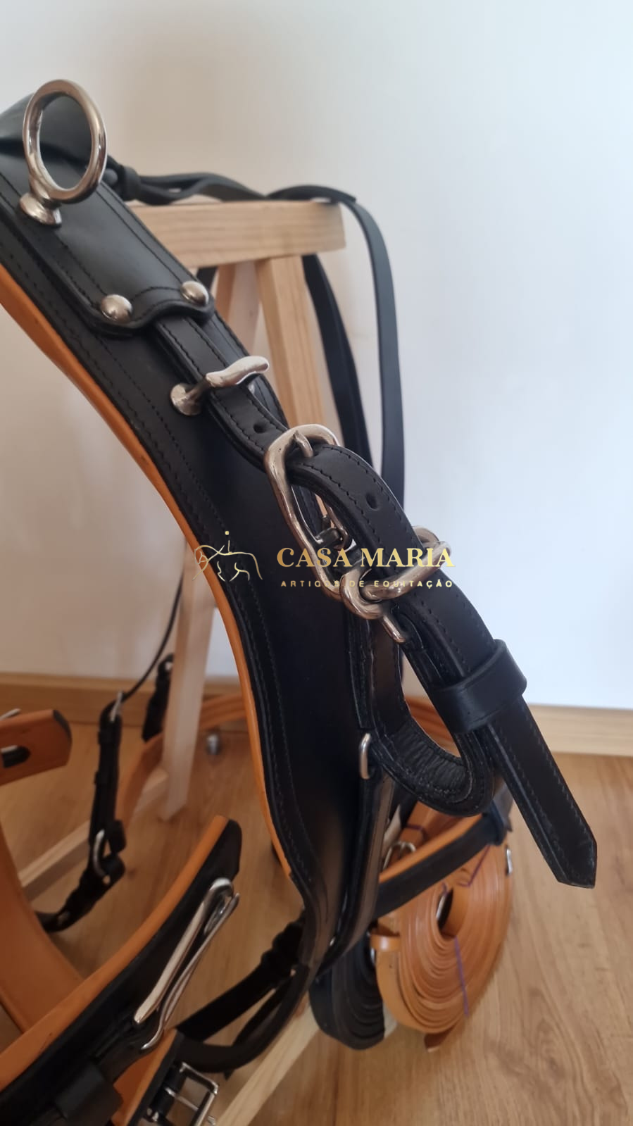 Peça de arreio em couro preto com fivelas metálicas pendurada em estrutura de madeira