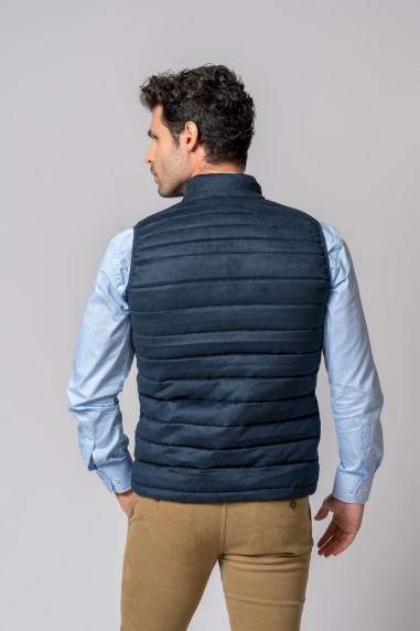 Homem de costas com colete azul escuro acolchoado, camisa azul clara e calças castanhas