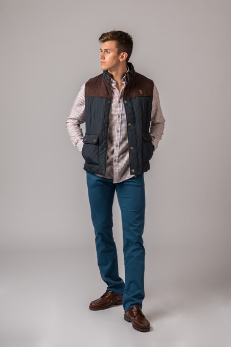 Homem com colete acolchoado azul escuro, camisa xadrez, calças azuis e sapatos castanhos num fundo cinzento