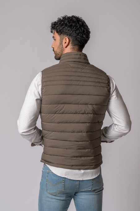 Colete castanho acolchoado visto de costas em modelo masculino com camisa branca e jeans azuis