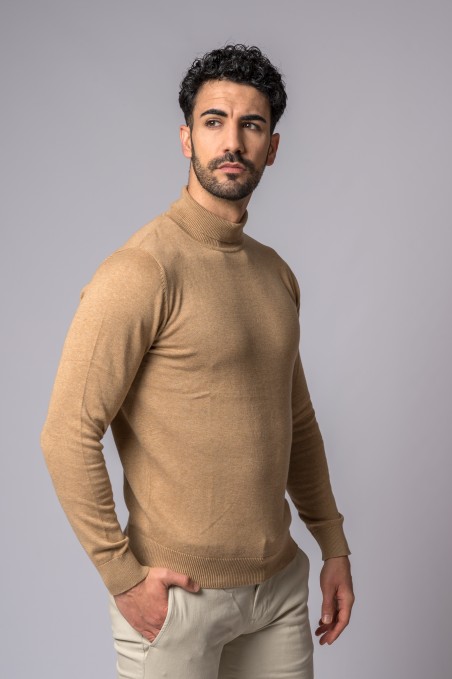 Homem com camisola de gola alta castanha clara e calças bege claras, fundo cinza liso.