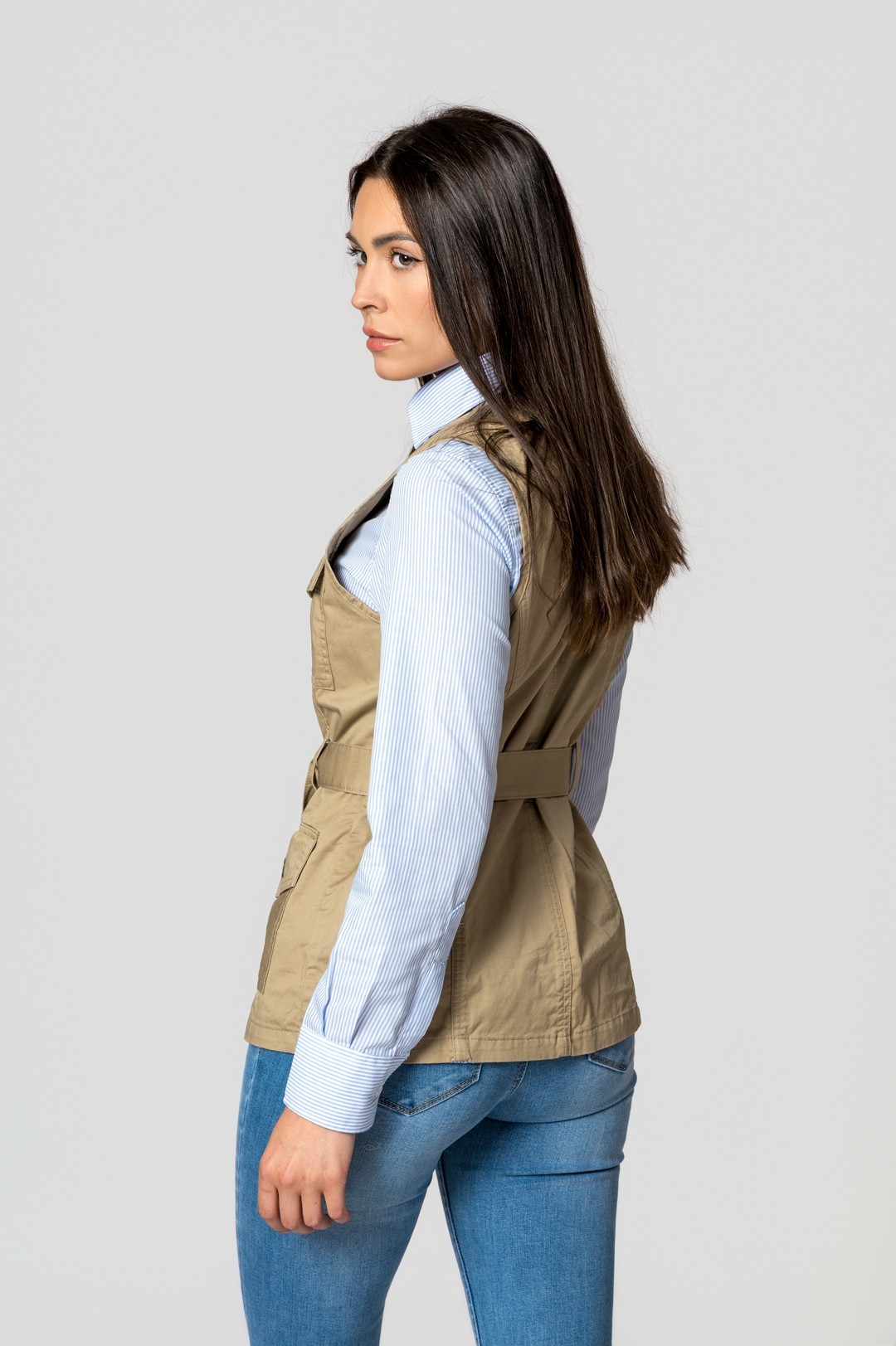 Mulher de perfil com camisa azul de riscas brancas, colete bege e jeans azuis.