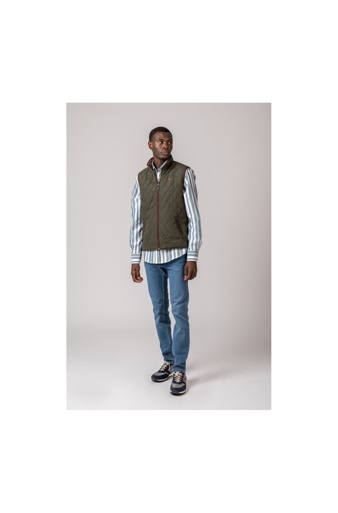 Homem vestido com camisa às riscas, colete verde matelassê, jeans azuis e sapatos desportivos.