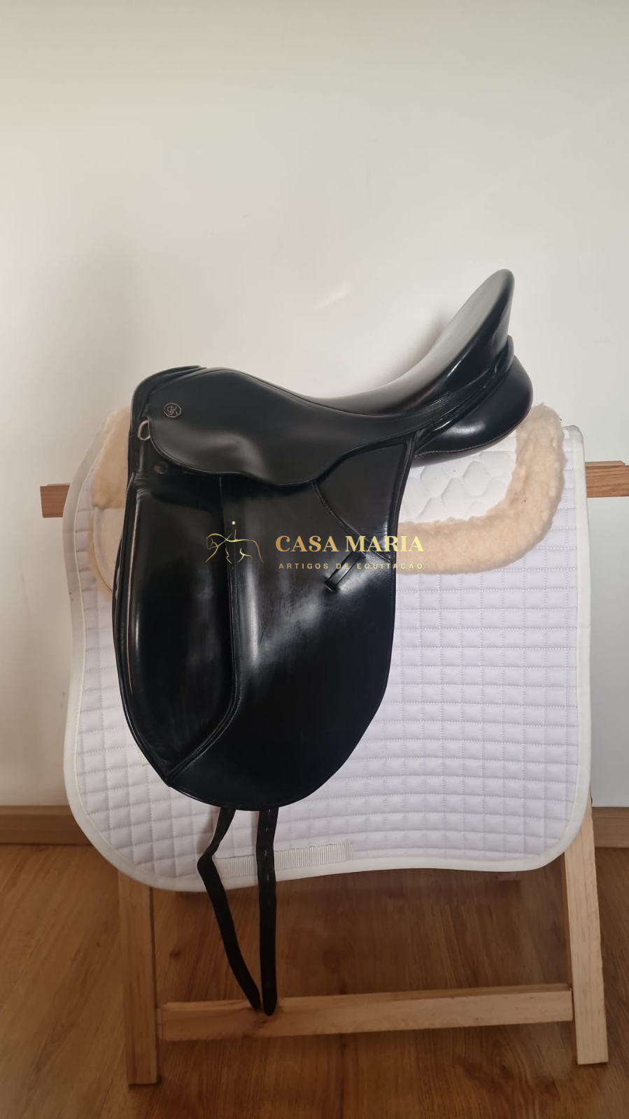 Sela de equitação preta em couro sobre manta branca acolchoada em suporte de madeira.