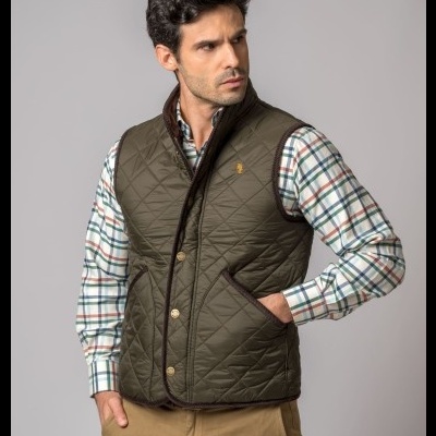 Colete masculino verde-oliva acolchoado com botões dourados sobre camisa xadrez e calças castanho claro