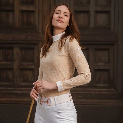 Mulher com blusa bege com padrão floral e calças brancas com cinto branco junto a porta de madeira