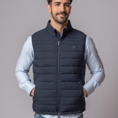 Homem a usar colete acolchoado azul escuro com camisa azul clara e jeans em fundo cinzento