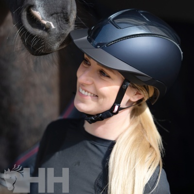 Mulher loira com capacete de equitação azul escuro sorrindo junto do focinho de um cavalo.