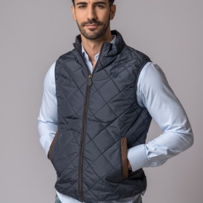 Colete acolchoado preto com bolsos castanhos usado sobre camisa azul com riscas finas e jeans azuis