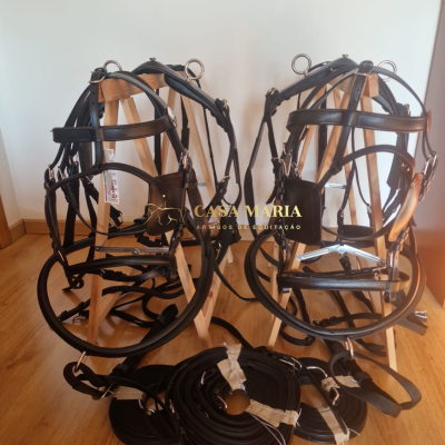 Dois arreios de cavalo em couro preto com rédeas, em cavaletes de madeira, sobre piso de madeira, fundo branco