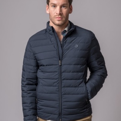 Homem com casaco acolchoado azul escuro e calças bege em fundo cinzento