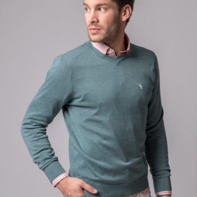 Homem com camisola verde-azulada, camisa xadrez rosa e branco e calças azul-acinzentadas