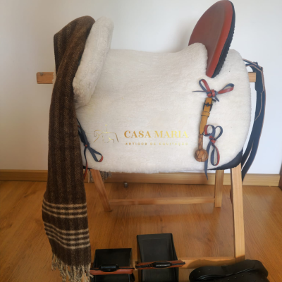 Sela de equitação branca com detalhes em couro castanho e texto dourado, manta xadrez, estribos e correias pretas em suporte de madeira