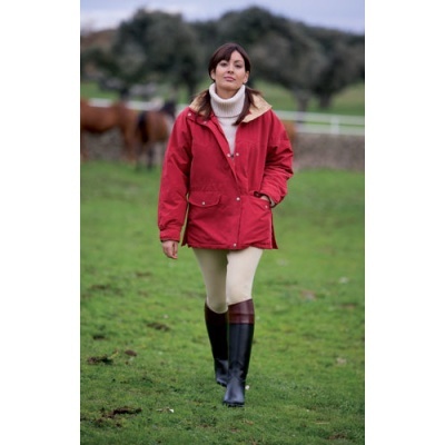 Mulher com casaco vermelho, calças bege e botas pretas a caminhar num campo com cavalos ao fundo.