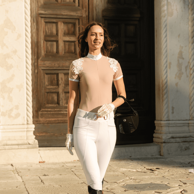 Mulher com roupa eqüestre bege e branca, luvas brancas e capacete preto numa rua com portas de madeira ao fundo.