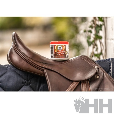 Sela de equitação em couro com frasco de bálsamo para couro em cima, sobre tecido azul escuro.