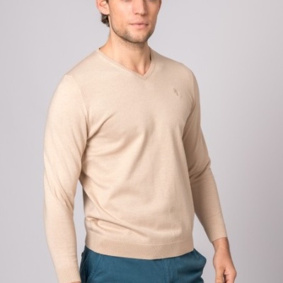 Homem com camisola de malha bege e calças azuis em fundo cinzento